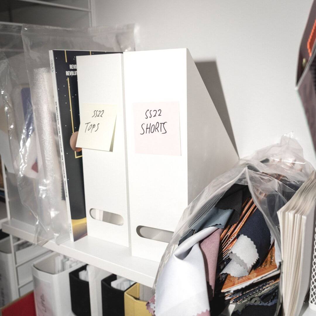 A white shelf with fabrics and two magazine folders labelled "SS22 Shorts" and "SS22 Tops" on post-it notes